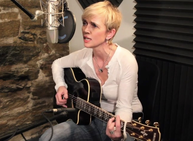 Girl in a recording room sitting on a chair with guitar singing to the mic
