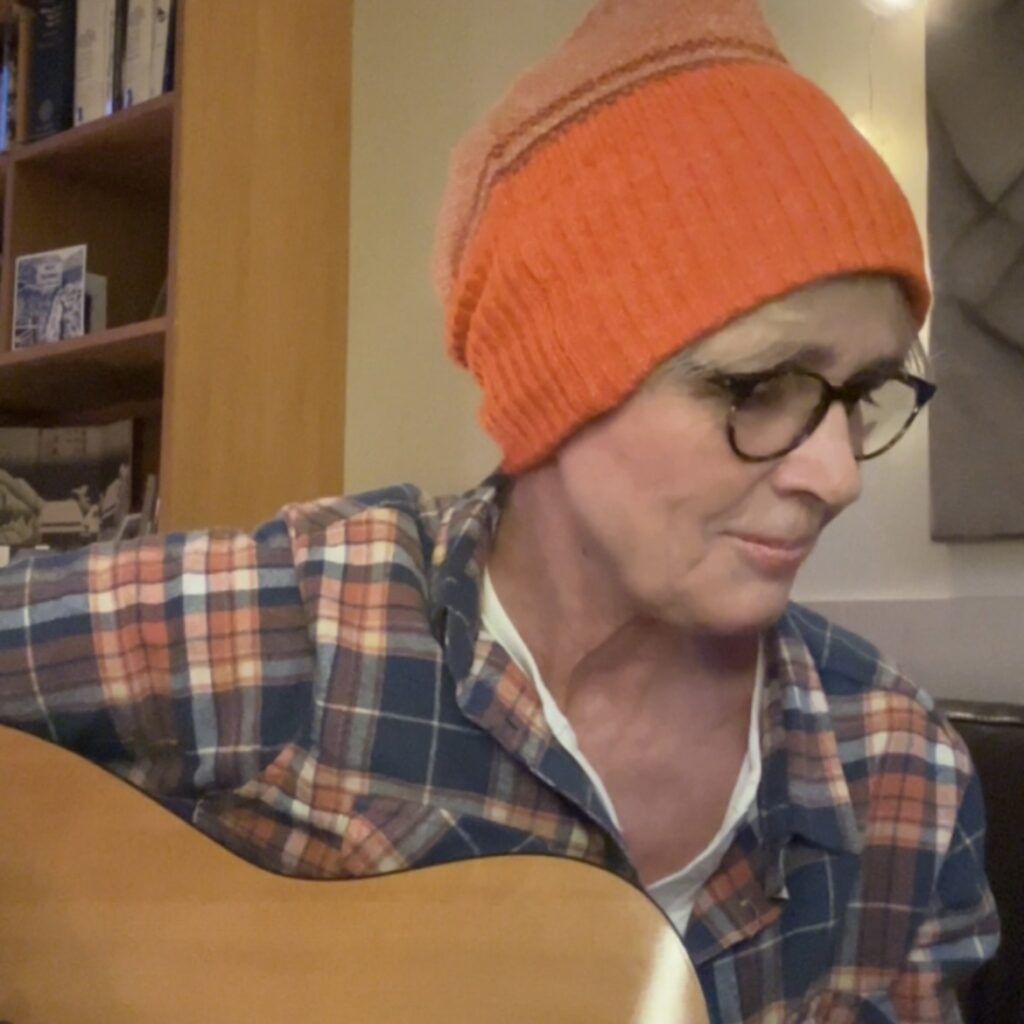 A woman with an orange beanie, hat, and orange check shirt is sitting on a sofa, looking down at her guitar and smiling.